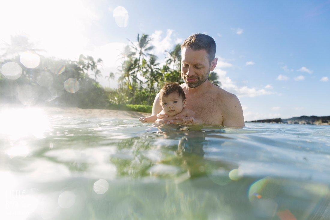 dad and newborn son in clear blue water of hawaii with sun flare