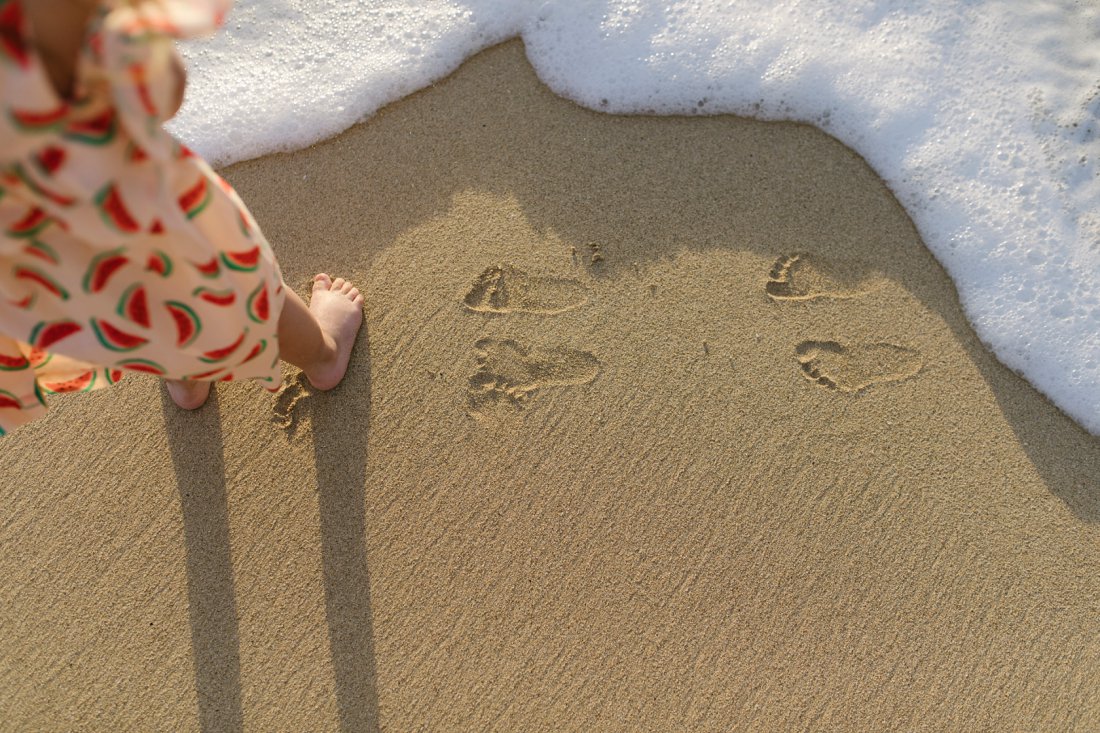 toddler feet with footprints in the sand and wave approaching