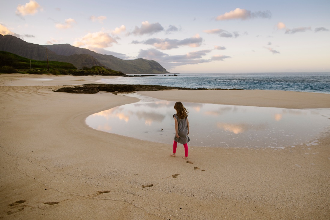 girl walking on the beach with clouds reflecting in a shallow pool and mountains in the background