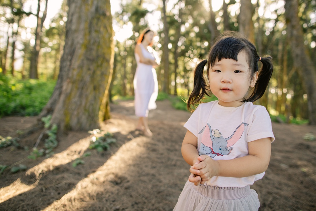 toddler girl in the forest with mom in the background by little bird photography