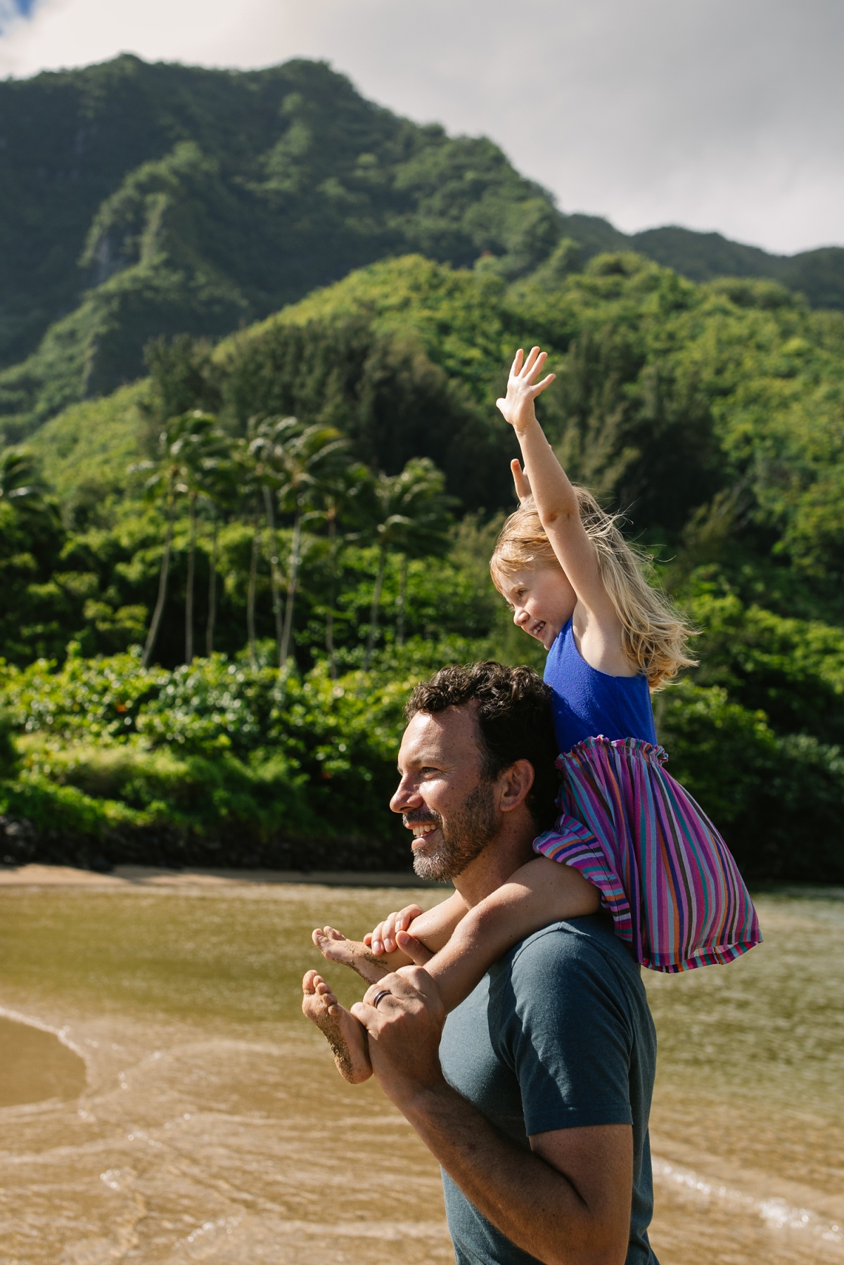 toddler riding on dads shoulders with mountains behind on windward oahu beach