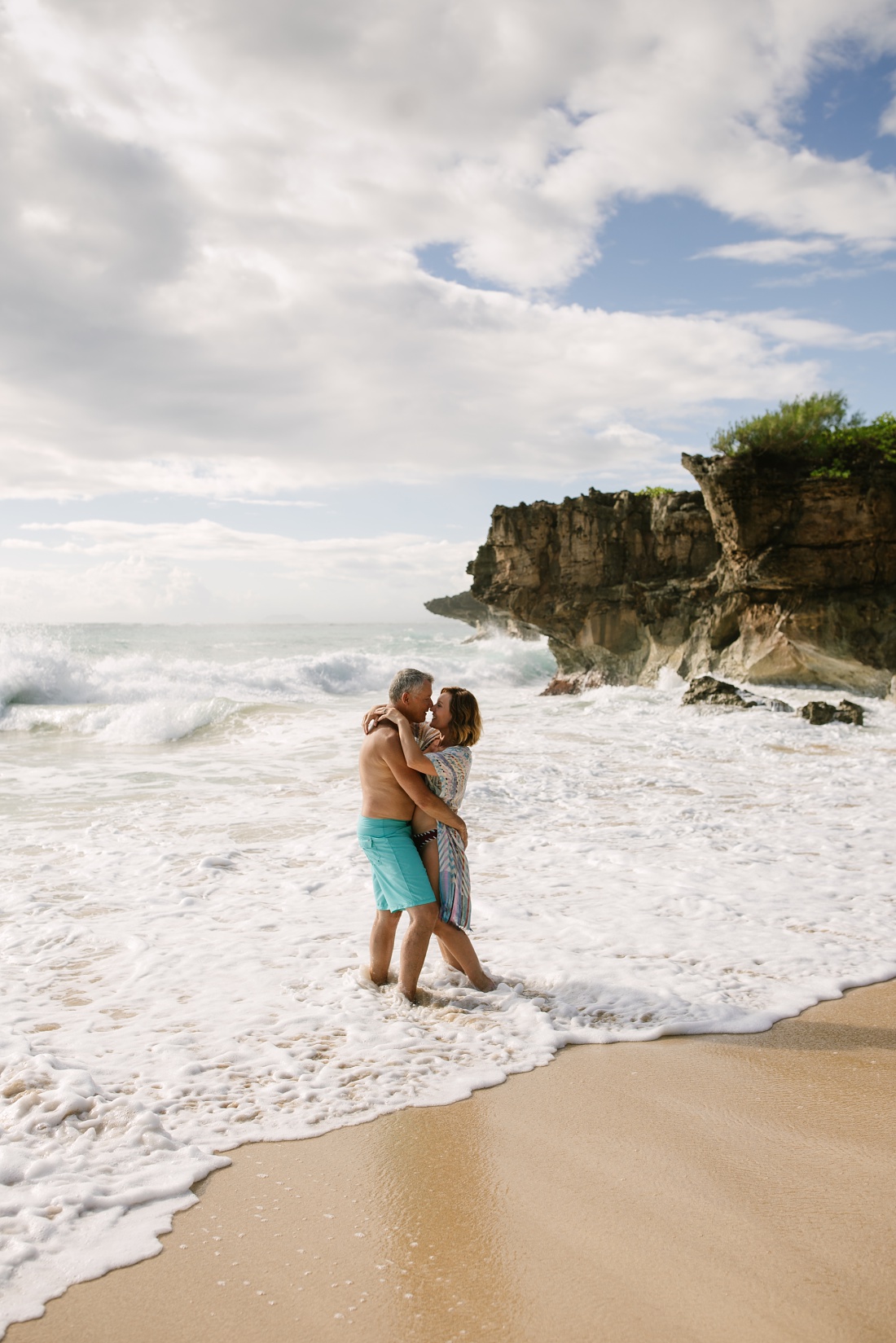 couple cuddles with waves splashing against cliffs in the background at pounders beach in laie