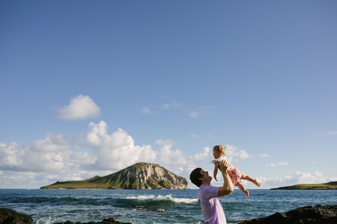 dad holds daughter in the air during a hawaii photo session with rabbit island behind
