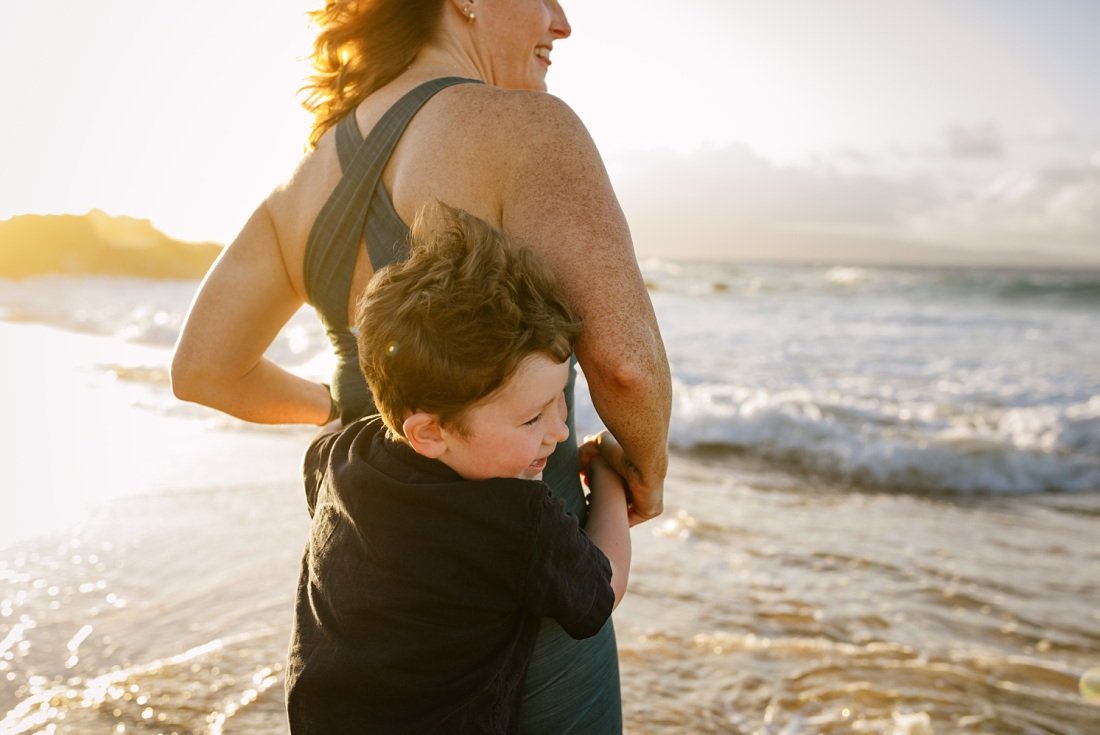 boy hiding from waves behind mom during family photos at kapalua ironwoods beach at sunset 