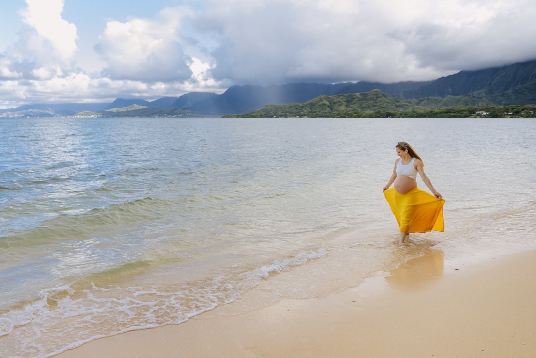 pregnant woman walking along the beach with mountains and rain behind oahu hawaii