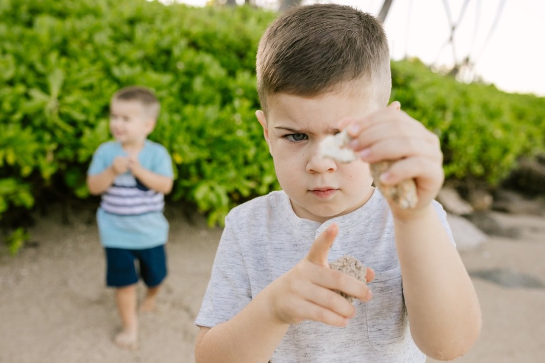 boy examining a piece of coral on the beach in hawaii with sibling behind