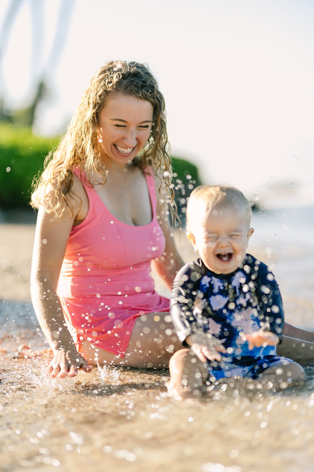 Family photographers in Oahu captures mom and baby playing in the water while he laughs with delight