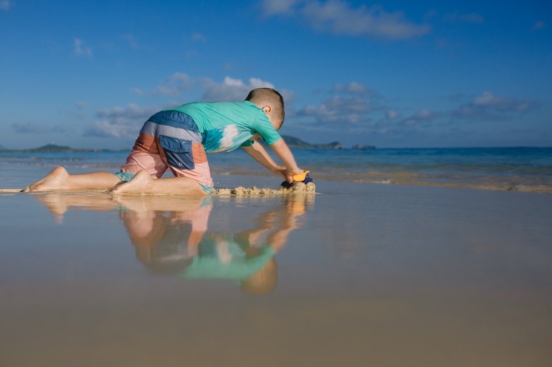 boy pushes a truck in the sand with his reflection clear against the blue water