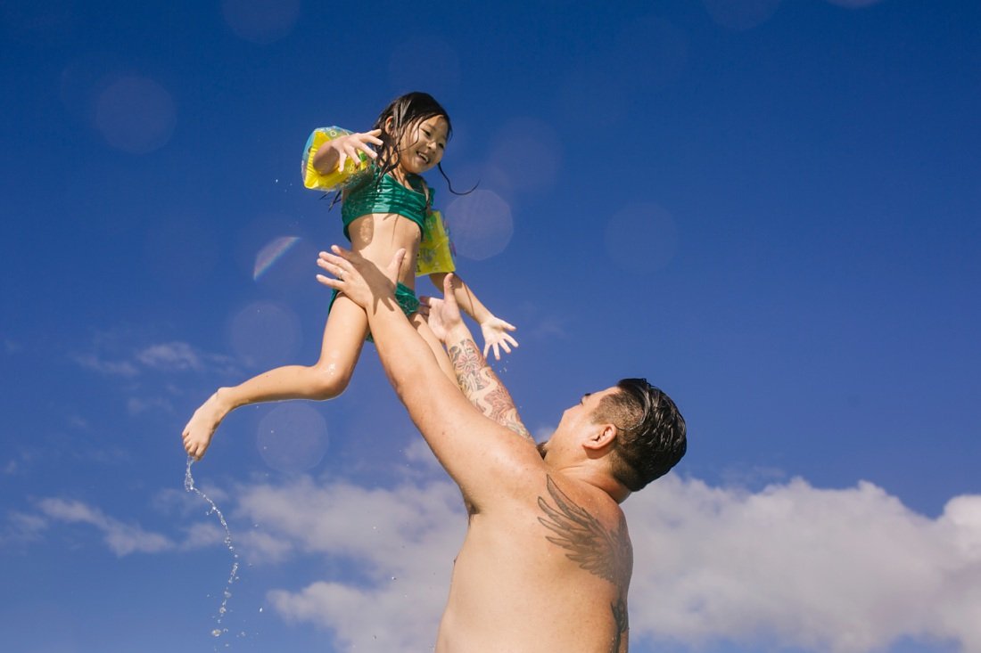 dad throwing daughter in the air against blue sky during fun family photo session in hawaii