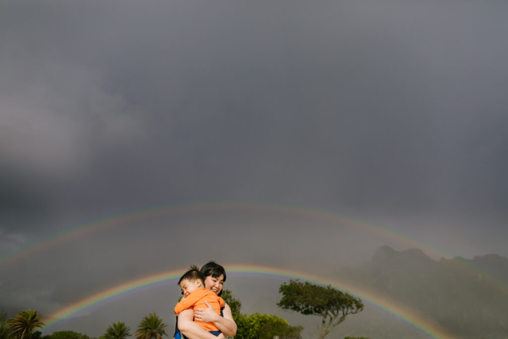 mom hugs her toddler son with a double rainbow against the koolau mountains behind during candid family photography Oahu